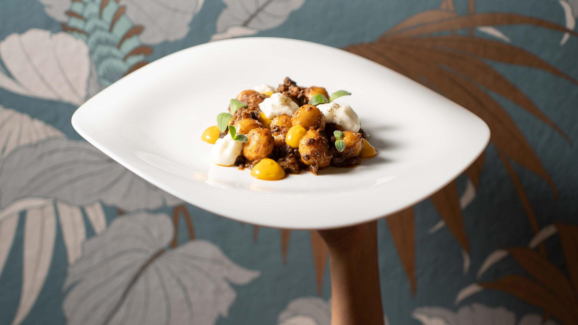 Piatto gourmet con gnocchi, crema gialla e decorazioni verdi su sfondo floreale.