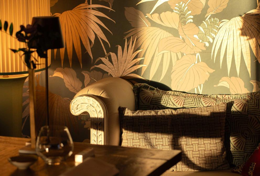 Soggiorno accogliente, luce calda, carta da parati tropicale, cuscini colorati, atmosfera rilassante.